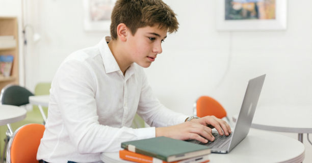A young boy wearing white long sleeves studies with a laptop in a classroom setting.