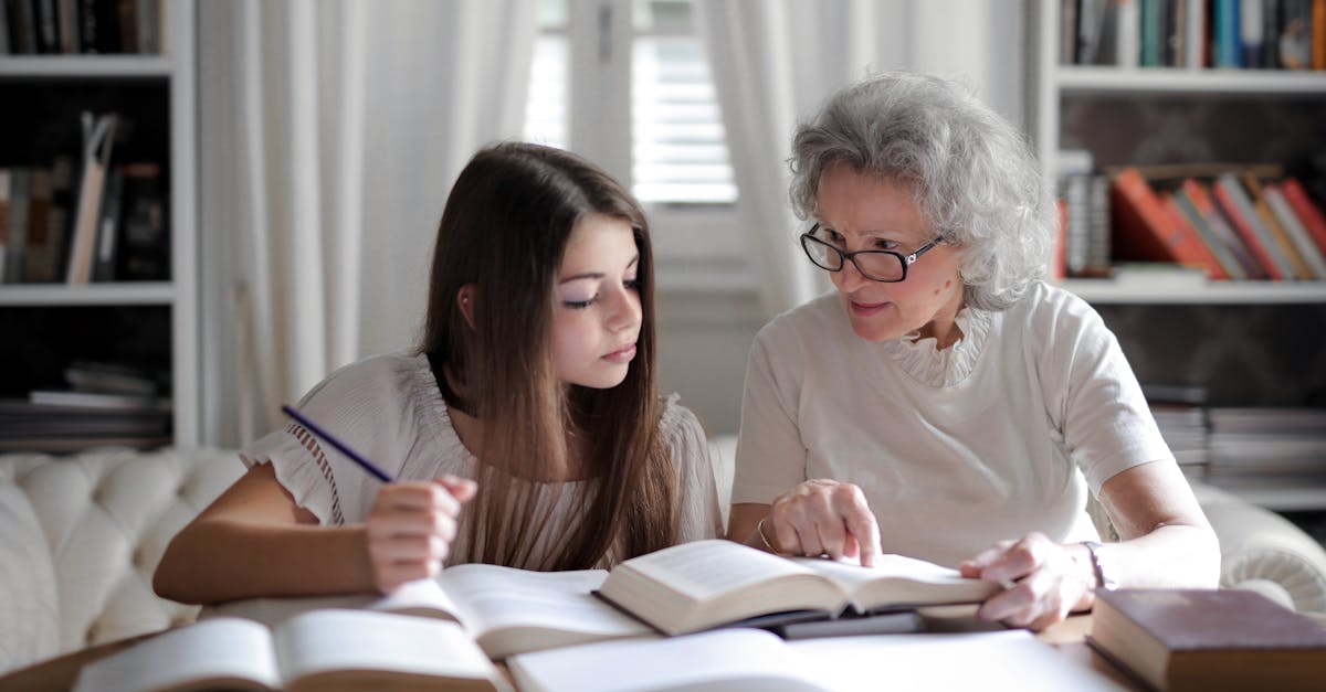 A grandmother helps her teenage granddaughter with homework, surrounded by books indoors.