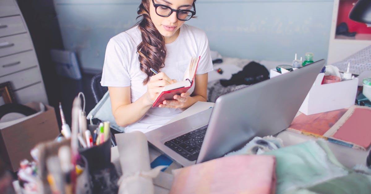 A young woman in glasses studies indoors with a laptop and notepad, writing notes.