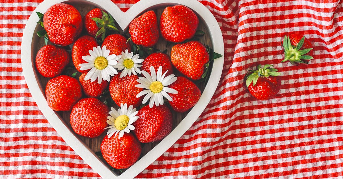 Fresh strawberries and daisies in a heart-shaped bowl on a red checkered cloth.