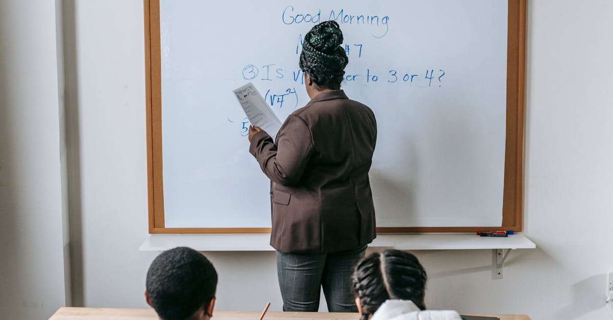 Back view unrecognizable pupils sitting at desk and listening to female teacher writing information on whiteboard