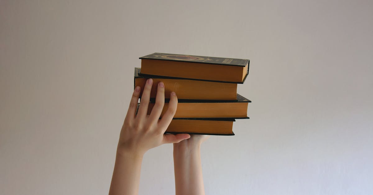 Hands reaching upwards holding a stack of hardbound books against a plain background.
