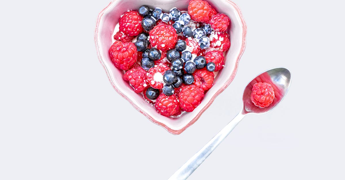Heart-shaped bowl filled with raspberries and blueberries, accompanied by a spoon on a white background.