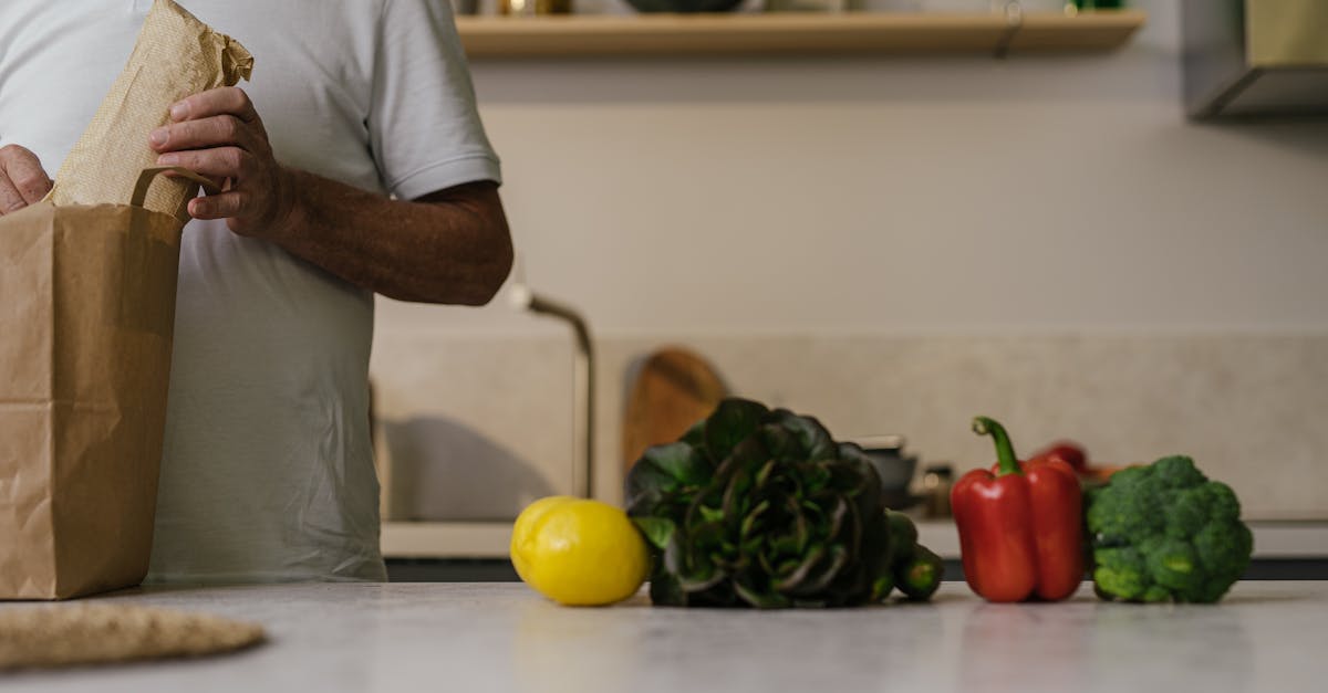 A man unpacks groceries from a paper bag on a kitchen counter with fresh vegetables.