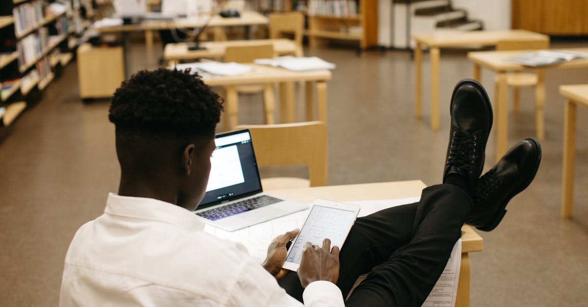 Black man studying in library with laptop and tablet, feet up on desk.