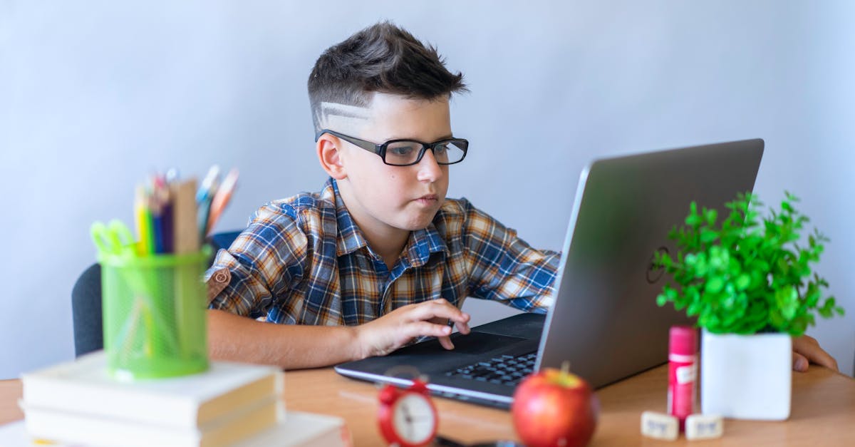 A young boy wearing glasses focused on using a laptop for online learning, surrounded by school supplies.