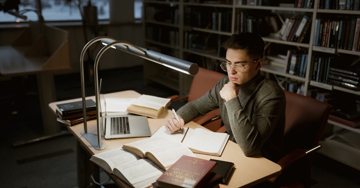 A young adult studying at a library desk with books and laptop under warm lighting.