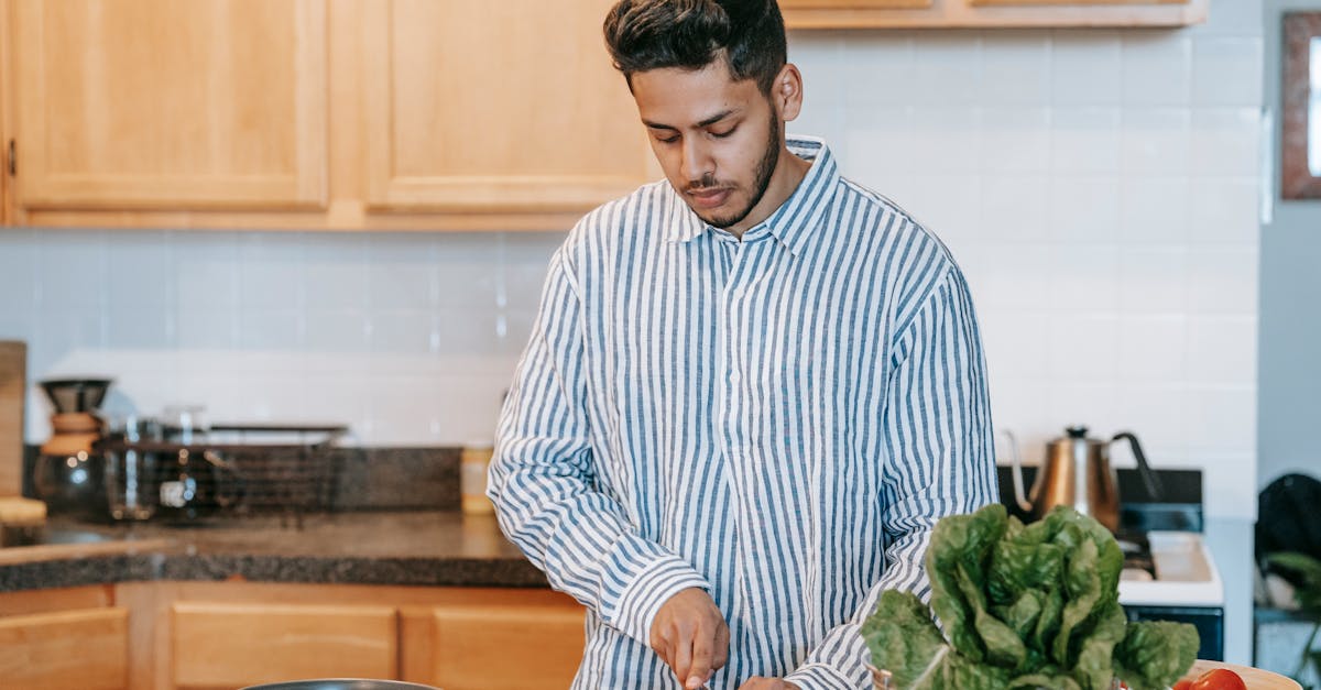 Bearded ethnic male in striped shirt cutting fresh tomato with knife against spinach leaves on table at home
