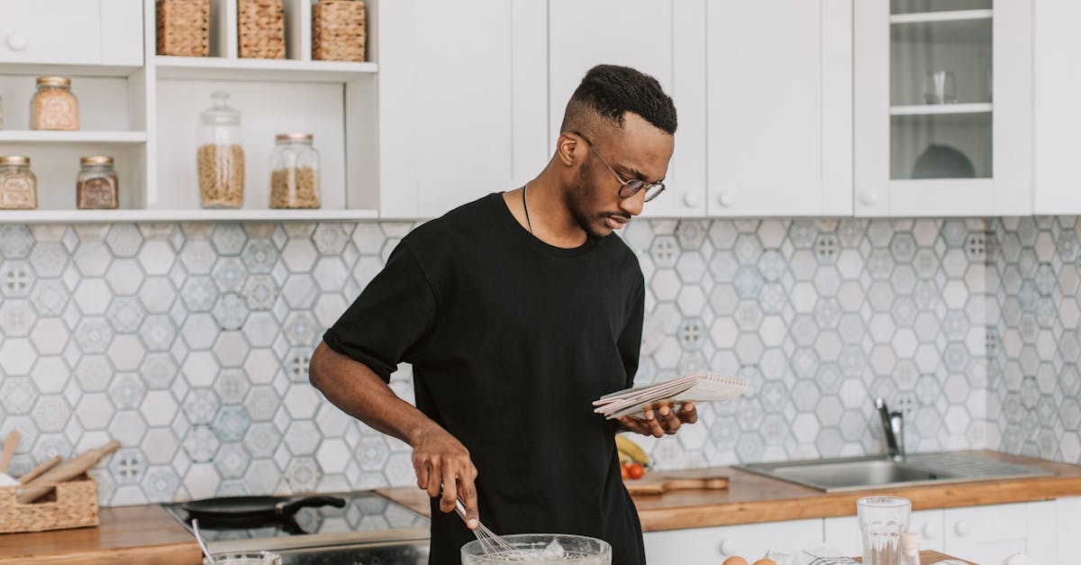 Black man preparing food in a stylish kitchen while reading a recipe book.