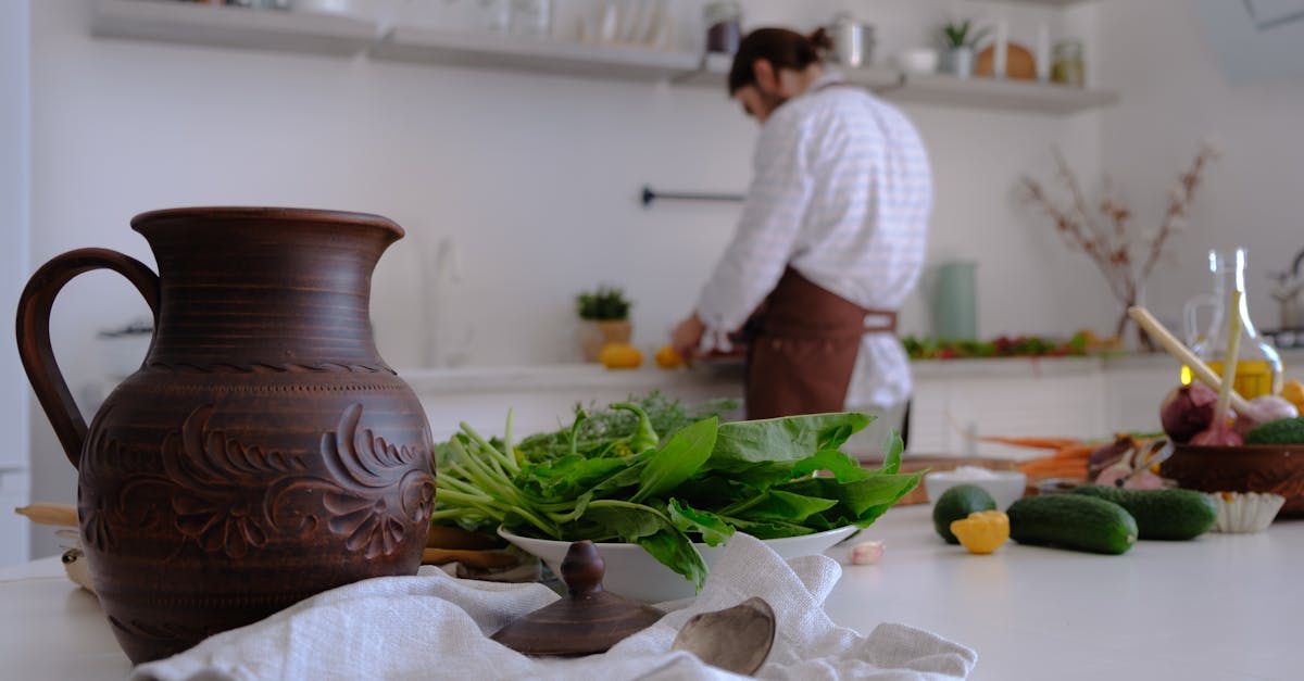 A man prepares fresh ingredients in a bright, rustic kitchen with a focus on a decorative jug and greens.