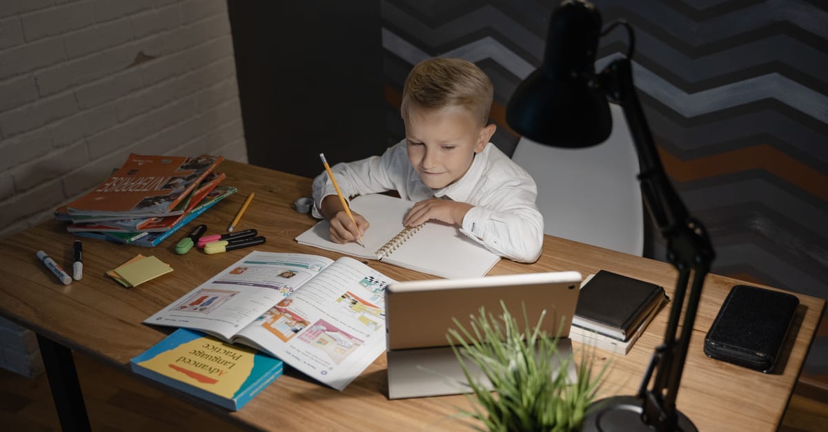 A young boy focused on studying at a wooden table with a tablet and books nearby under a desk lamp.