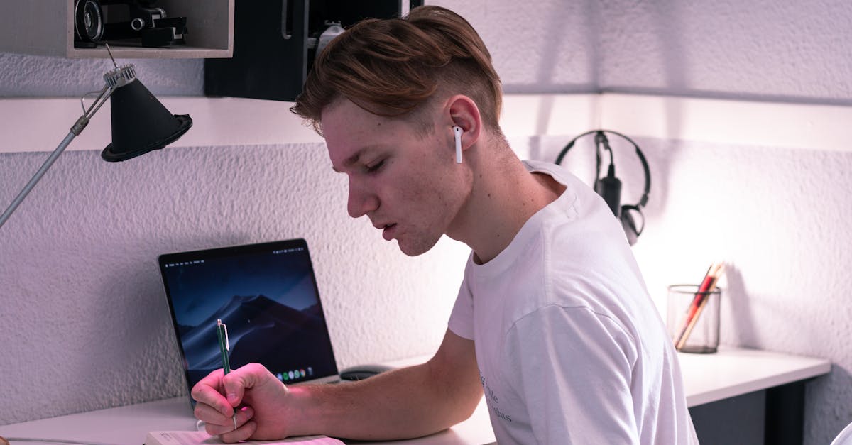 Side view of serious young man in TWS earbuds writing in notebook while studying on netbook at desk