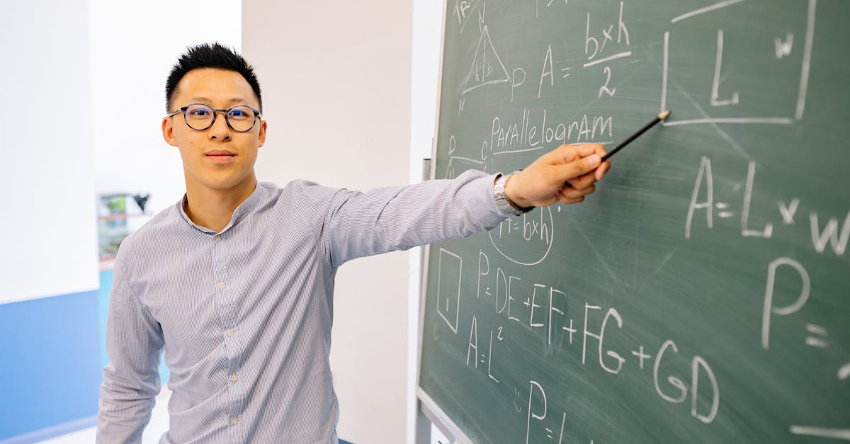 An Asian male teacher wearing eyeglasses points confidently at a blackboard filled with math equations in a classroom setting.
