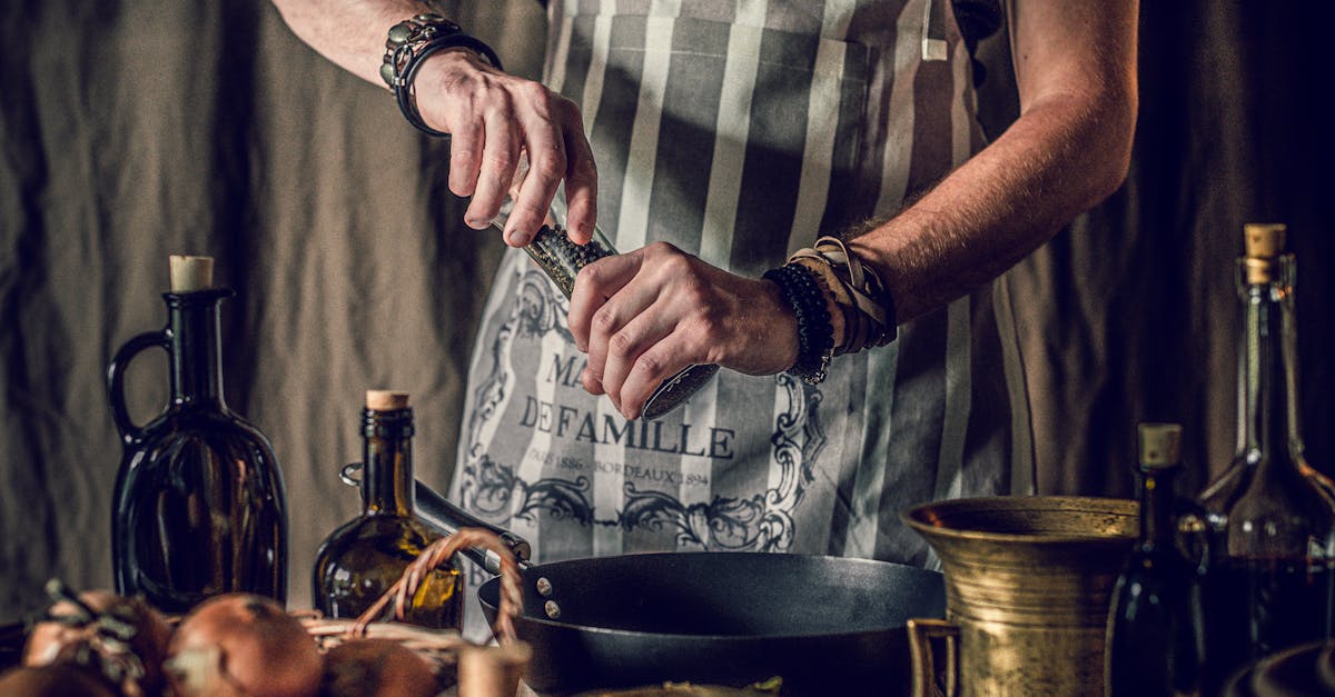 Crop faceless man in apron adding pepper in pan while cooking traditional dish standing at table with assorted ingredient and oil bottles in rustic kitchen