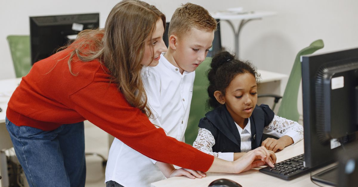 Young teacher helps students with a computer task in a classroom setting.