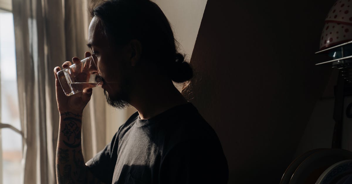 A man standing indoors, drinking water from a glass near a window, reflecting in soft light.