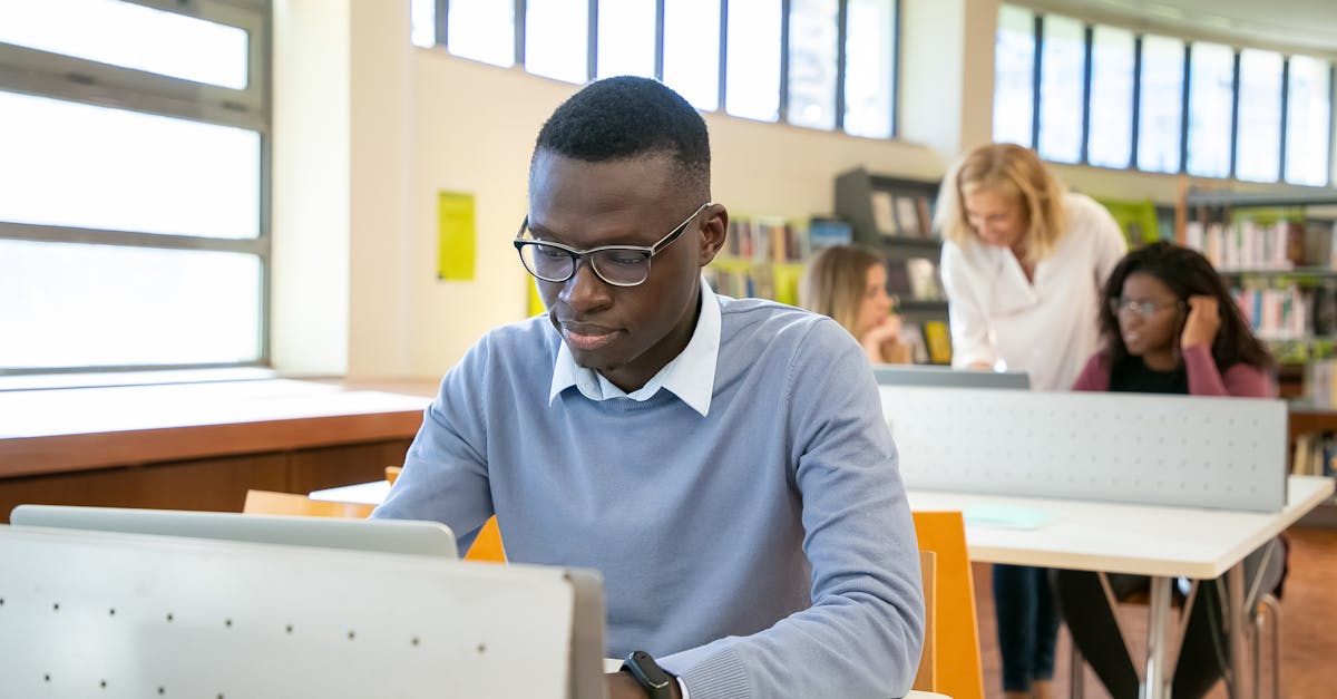 Concentrated young black male student in casual clothes and eyeglasses using laptop sitting at desk in classroom with multiethnic group of mates in college