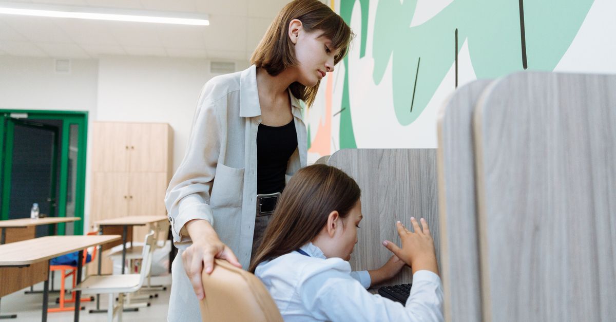 A teacher assists a student working on a computer in a bright, modern classroom setting.
