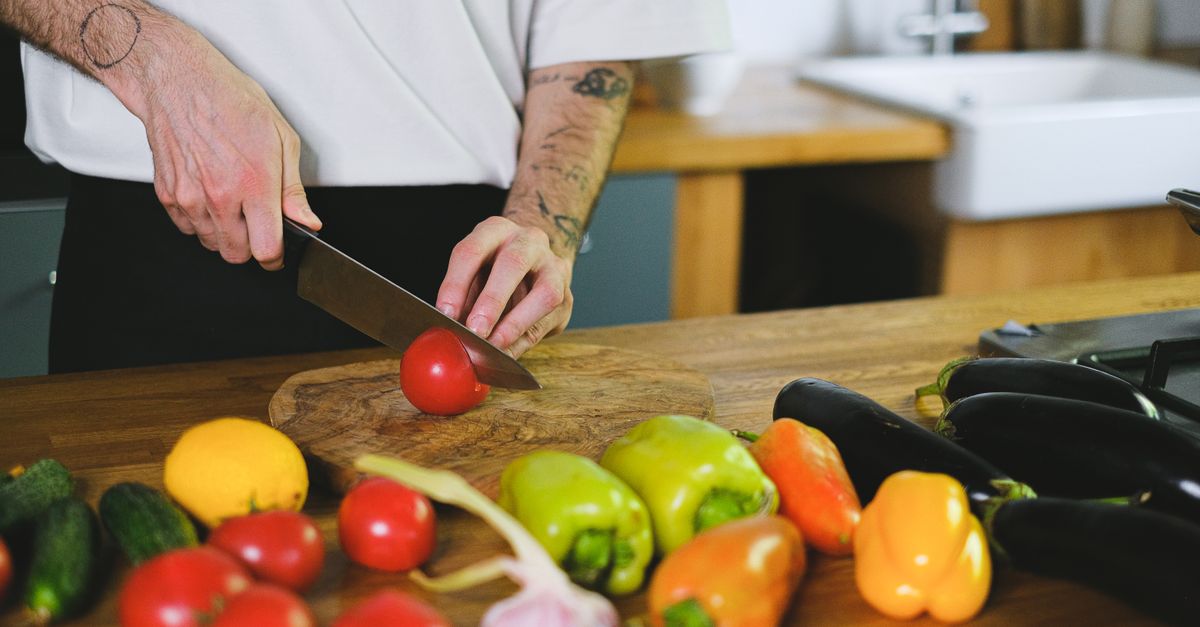 A person slicing tomatoes on a wooden board among fresh vegetables in a kitchen setting.