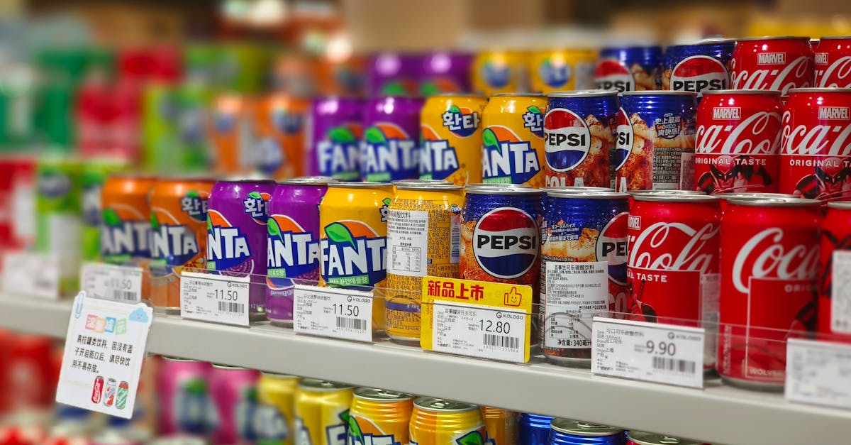 Colorful soda cans including Fanta, Pepsi, and Coca-Cola arranged on supermarket shelves, price tags visible.