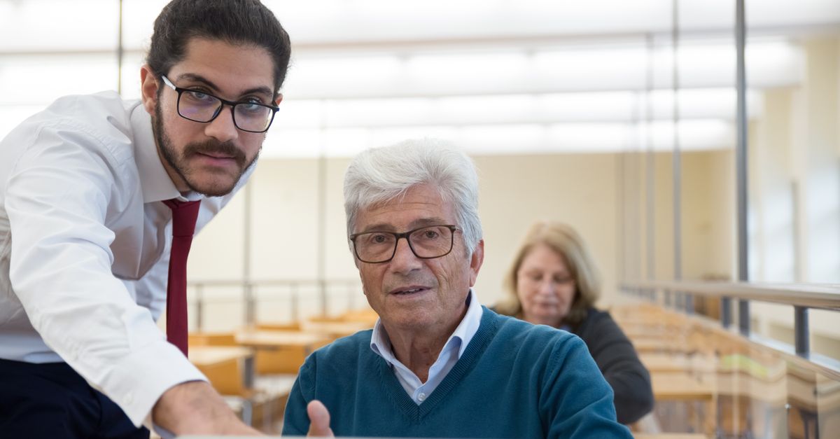 An elderly man learning computer skills with a teacher in a classroom setting, showcasing education through technology.