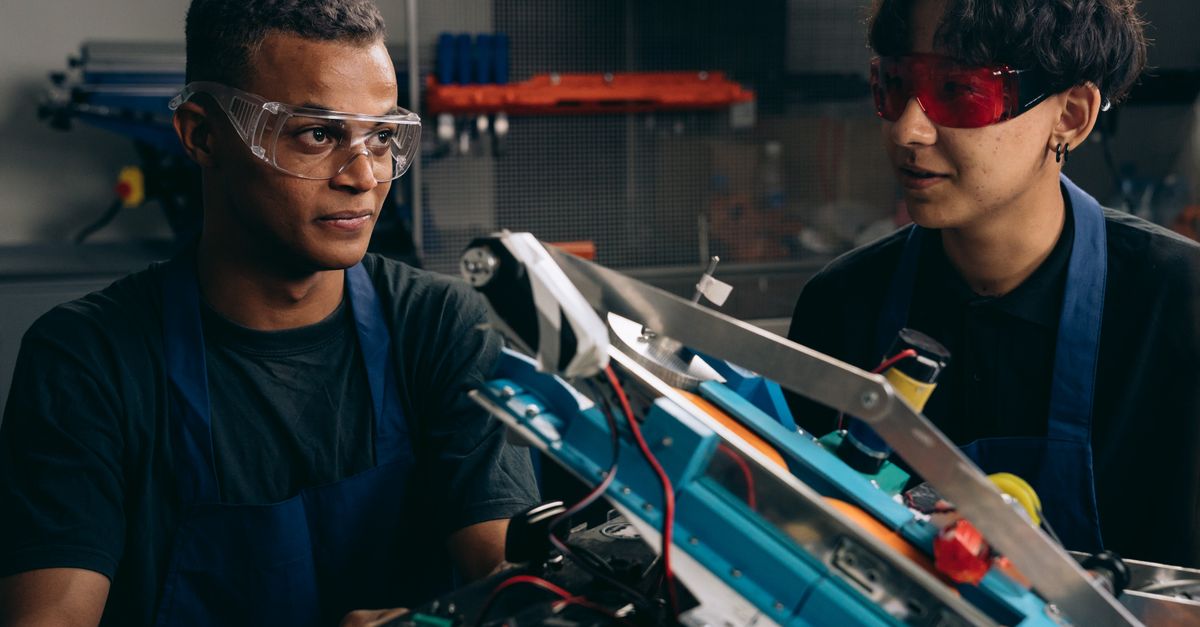 Two young men focused on assembling a robotic prototype, wearing safety glasses and aprons.