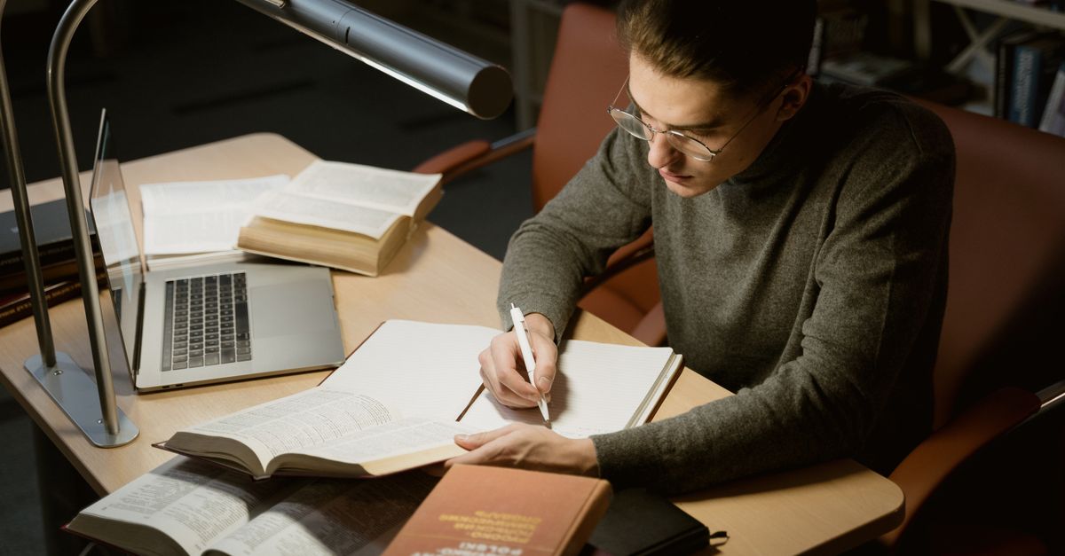 A young adult studying with books and a laptop, focused on taking notes at a desk under a lamp.
