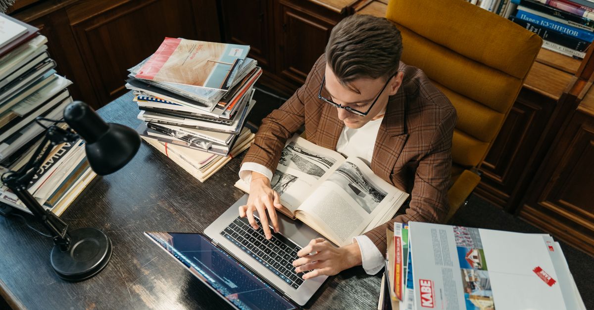 A young man studies in a cozy library setting, using a laptop and books at a wooden desk.