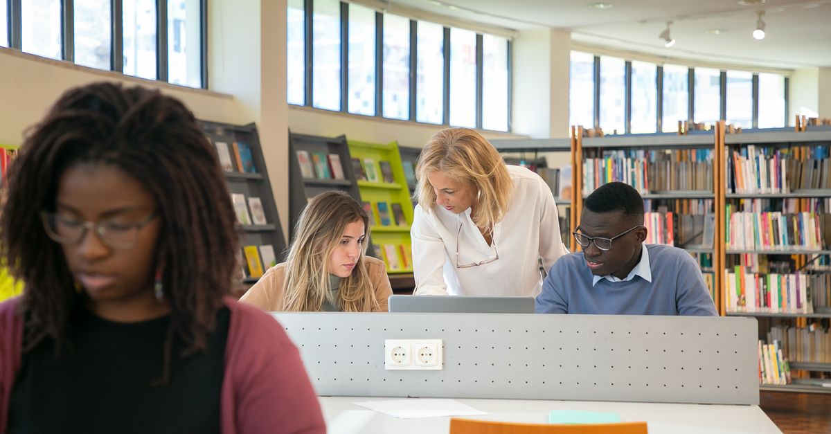 Elegant middle aged female library worker helping to young multiracial students to find information during work on laptop