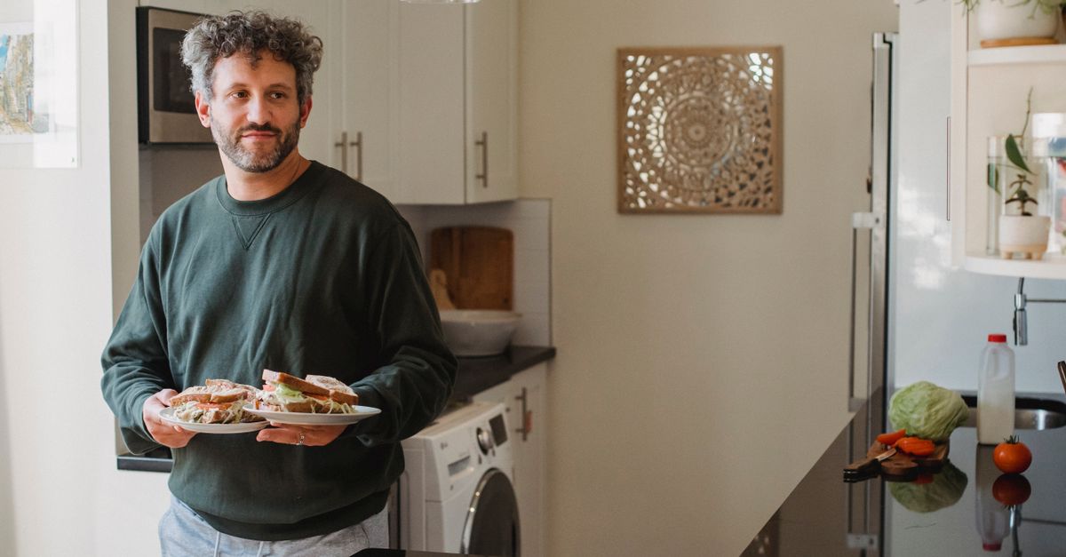 Tranquil man with beard and curly hair in casual clothes carrying plates with delicious sandwiches in cozy kitchen at home in daylight