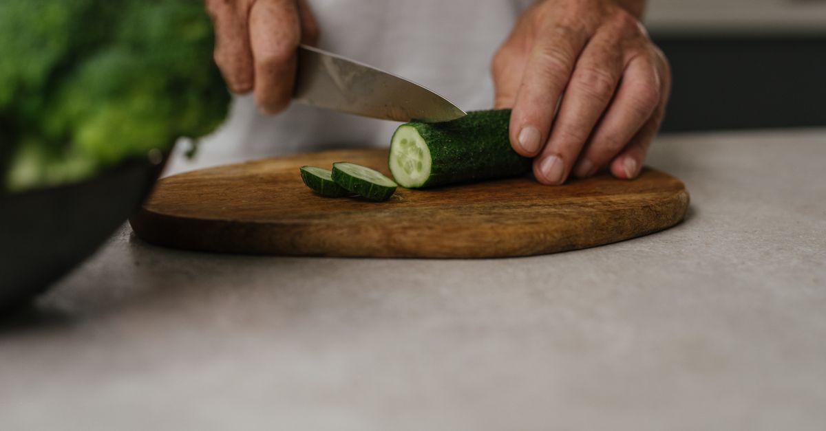 Close-up of hands slicing cucumber on wooden board in kitchen setting.