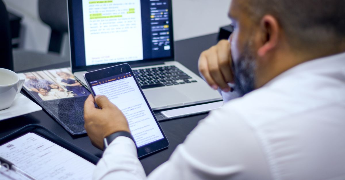 Man in white shirt working on a laptop and tablet at an office desk.