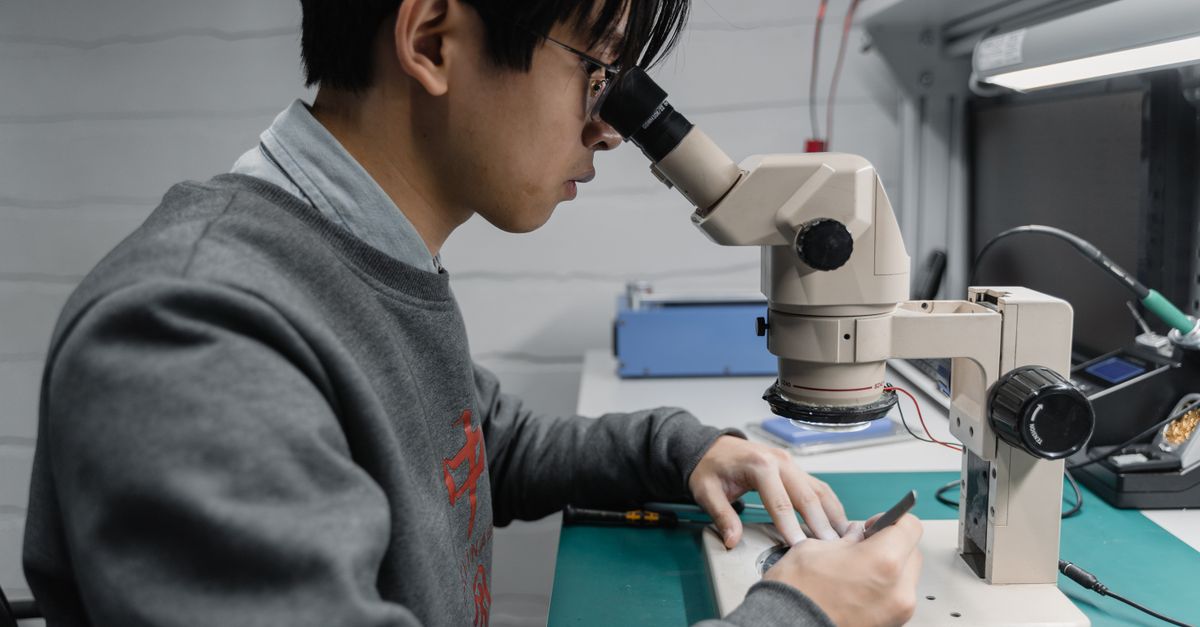 Young male technician working with a microscope in a laboratory setting, focused on electronics.