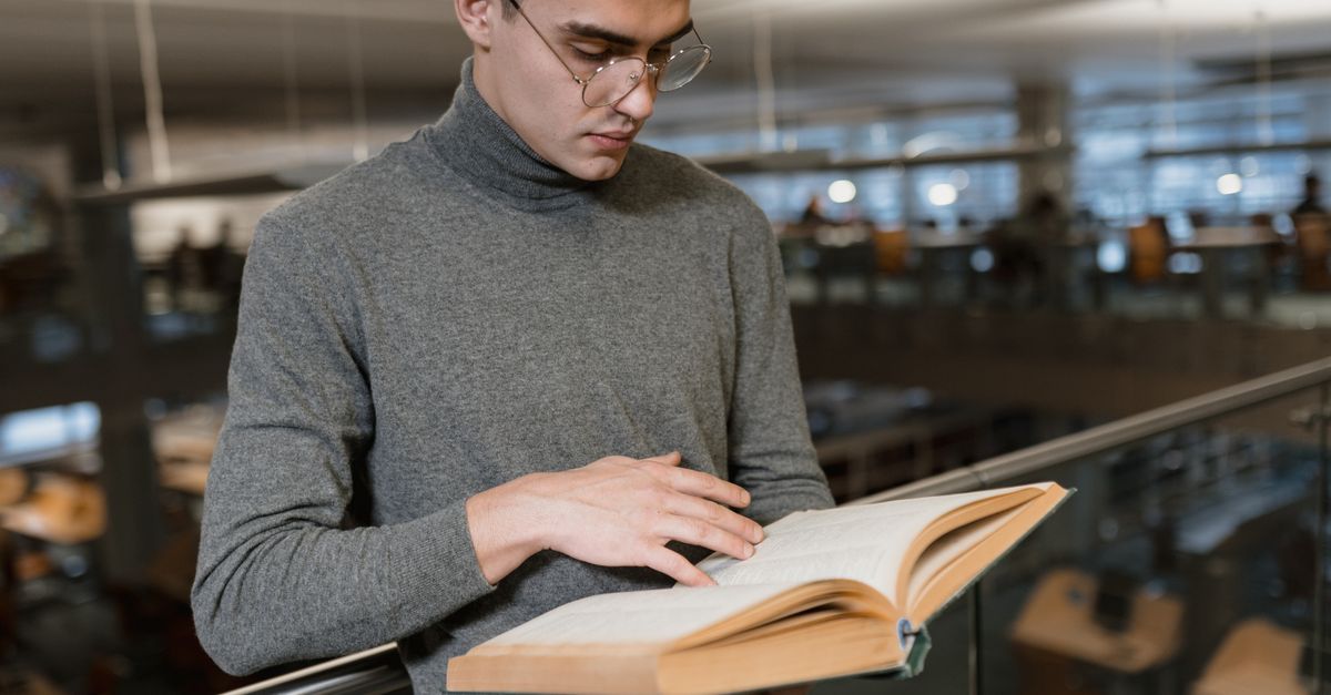 Adult man in gray turtleneck reading a book inside a modern library.